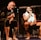 Two individuals are seated on a stage, each holding a microphone and smiling. They are in a sports-related setting with one wearing a casual black outfit and the other in sportswear, with a towel around his neck. The background includes elements of a fighting or martial arts cage.