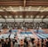 A large indoor arena with a high ceiling, filled with an audience seated on bleachers. In the center, two fighters are engaged in a martial arts competition on blue and red mats. The scene is captured through a mesh fence, adding a textured foreground.