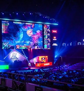 A large esports arena featuring a massive screen displaying a video game, surrounded by sponsor logos and team names. The auditorium is filled with spectators watching the gaming event. Neon blue and purple lights illuminate the stage, creating a vibrant atmosphere.