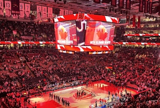 An indoor sports arena filled with spectators and a basketball court illuminated by red lighting. The scoreboards and video screens display promotional content, with numerous banners and championship flags hanging from the ceiling. Players and officials are gathered on the court, possibly in preparation for a game or event.