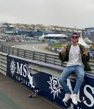 A person in casual attire sits on a barrier beside a racetrack, making peace signs with both hands. In the background, there is a stadium, a large crowd, promotional banners, and a screen displaying an advertisement. The scene has an overcast sky, and the setting appears to be a motorsport event.