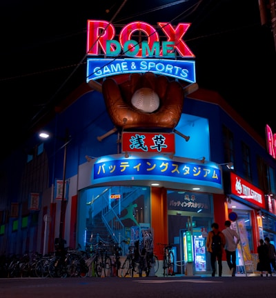 A vibrant, neon-lit facade of a gaming and sports establishment at night, featuring a large glove holding a baseball above the entrance. The building is adorned with colorful signs in both English and Japanese, with various advertisements and an illuminated sign that says 'ROX DOME'. Several people are walking towards the entrance, and bicycles are parked along the side.