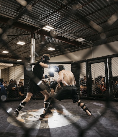 Two fighters engage in a mixed martial arts match within a caged ring. They are wearing protective gear, including gloves and shin guards, and are surrounded by spectators watching intently. Harsh lighting from above casts shadows on the fighters and the mat.
