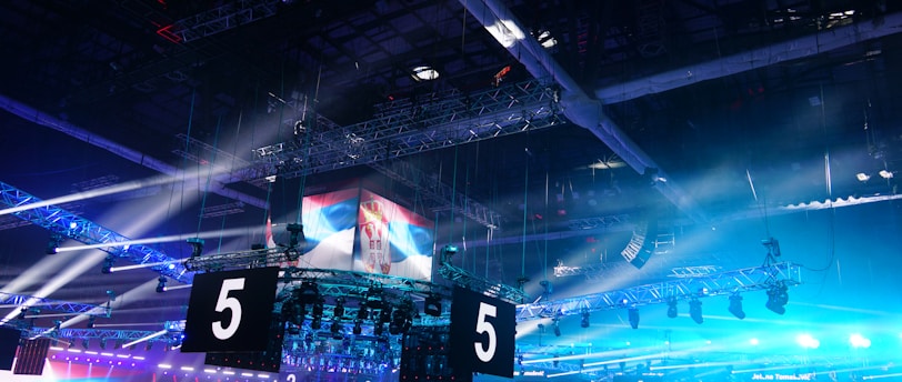 A large indoor arena is equipped with advanced lighting and audio systems. Suspended above the space are numerous digital displays showcasing numbers. The environment is filled with blue illumination and spotlights creating a vibrant atmosphere.