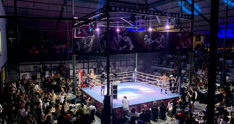 A crowded indoor arena with a boxing ring at the center, surrounded by an audience seated at tables. The scene is illuminated by overhead lights, casting a vibrant glow over the ring where two fighters are preparing for a match. Spectators are engaged, capturing the event with cameras and enjoying the lively atmosphere.