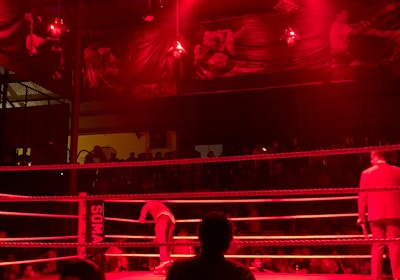 A wrestling ring is illuminated with red lighting, creating a dramatic atmosphere. Several people are visible around the ring, with silhouetted spectators in the foreground. Banners or art adorn the upper walls, and the venue has an industrial ceiling with exposed beams.