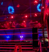A brightly lit boxing or wrestling ring with vivid red and blue lights. Banners with images of fighters hang from the ceiling. The ropes of the ring are prominently visible, and a stool is placed inside the ring. The arena is filled with an audience, silhouetted against the colorful lighting.