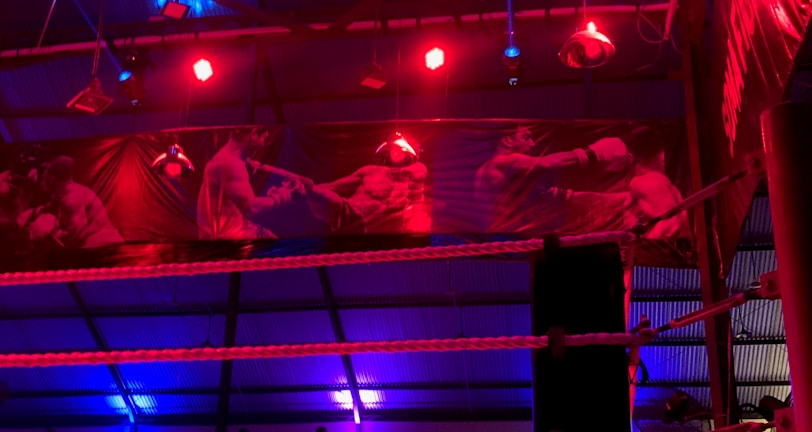 A brightly lit boxing or wrestling ring with vivid red and blue lights. Banners with images of fighters hang from the ceiling. The ropes of the ring are prominently visible, and a stool is placed inside the ring. The arena is filled with an audience, silhouetted against the colorful lighting.